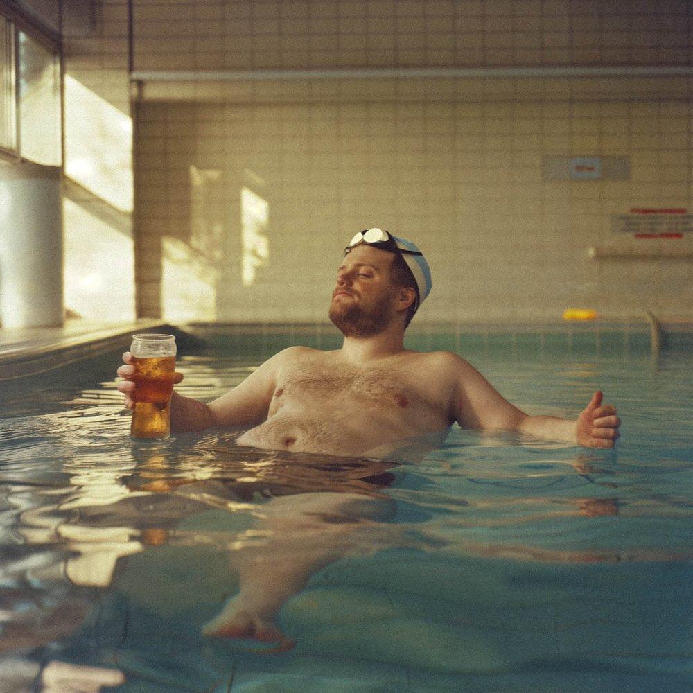 A man in a swimming pool, wearing a pair of goggles on his forehead, holding a glass of beer and giving a thumbs-up.