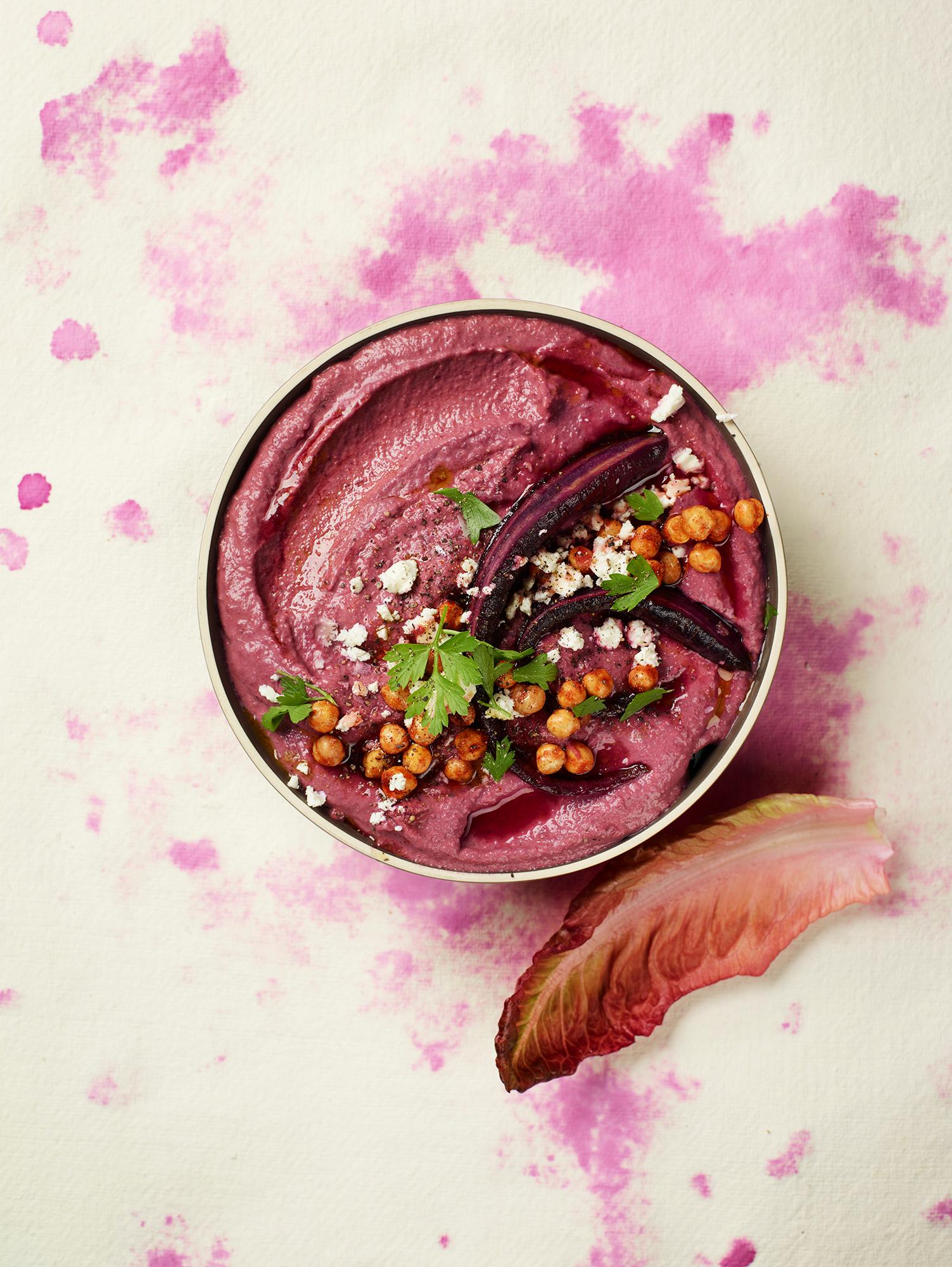 A bowl of vibrant, pink beet hummus, garnished with fresh herbs and crispy chickpeas, served in a rustic, wooden bowl on a natural, stone table.