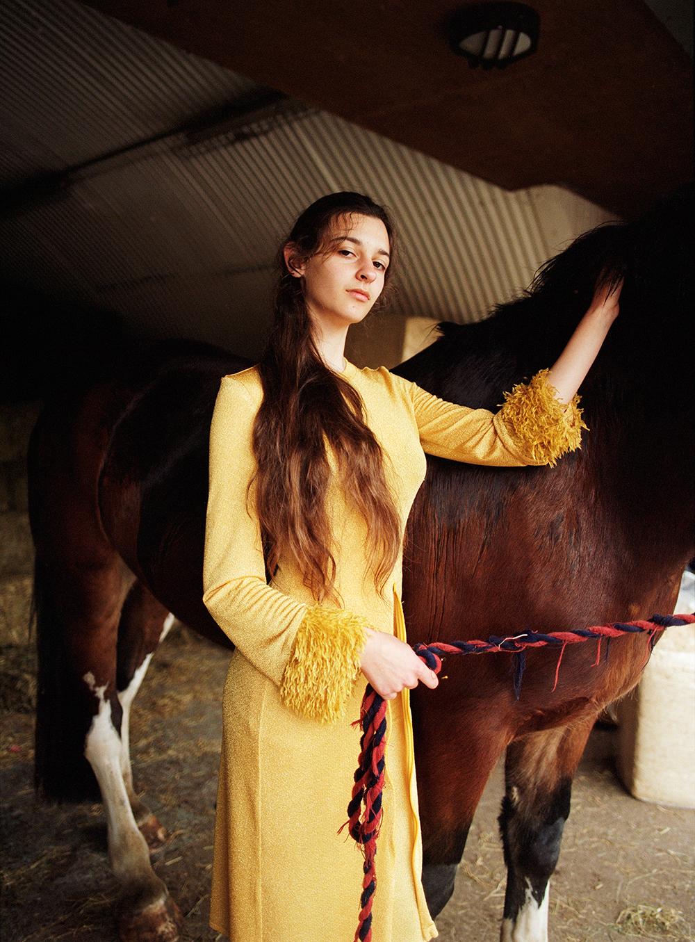 A woman in a yellow dress standing in a stable, holding a horse's halter and stroking its neck.