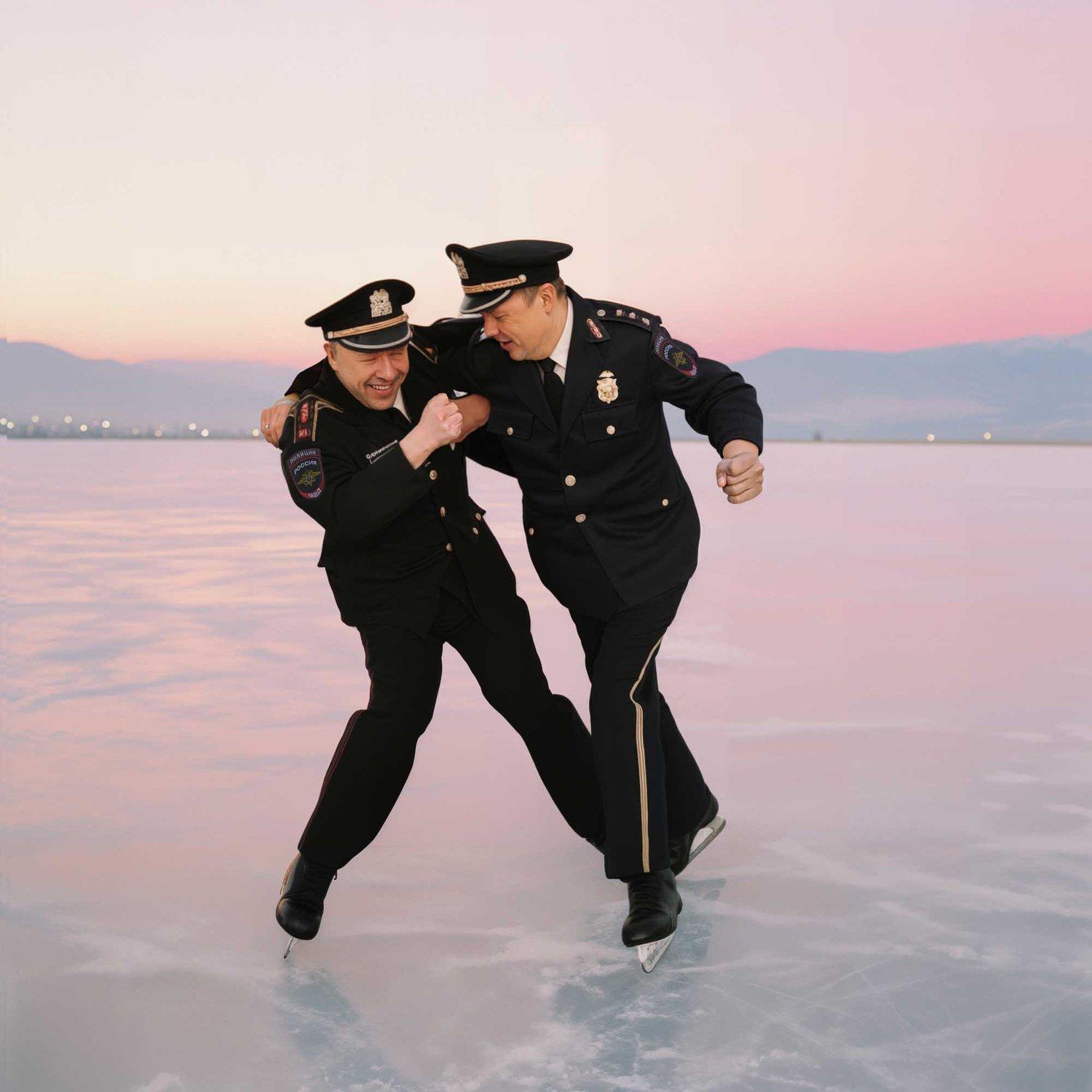 Two police officers in uniform are ice skating on a frozen lake, with a mountain range in the background.