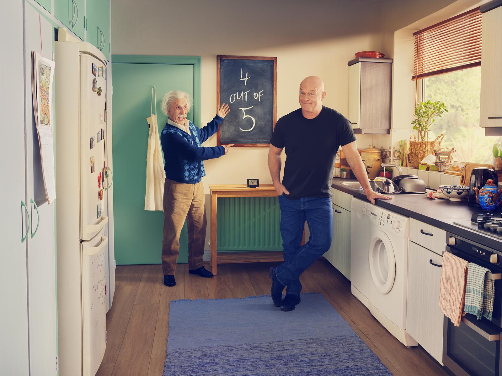 A man in a black t-shirt and jeans stands in a kitchen, with a man in a blue sweater and jeans standing next to a blackboard with the equation '4 out of 5' written on it.