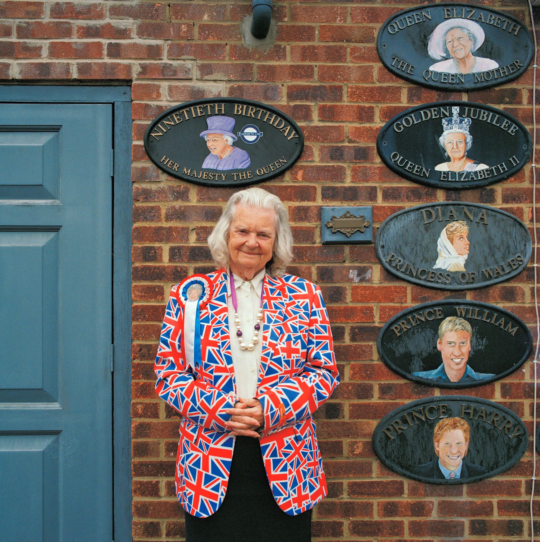 An elderly woman stands in front of a brick wall with commemorative plaques.