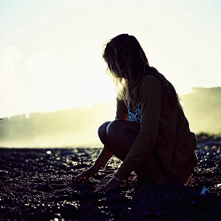 A woman is crouched on a beach, her long hair blowing in the wind as she reaches out to touch the sand.
