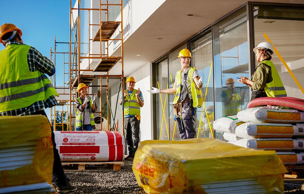 A group of construction workers in yellow hard hats and high-visibility vests are standing on a construction site, surrounded by building materials and scaffolding.