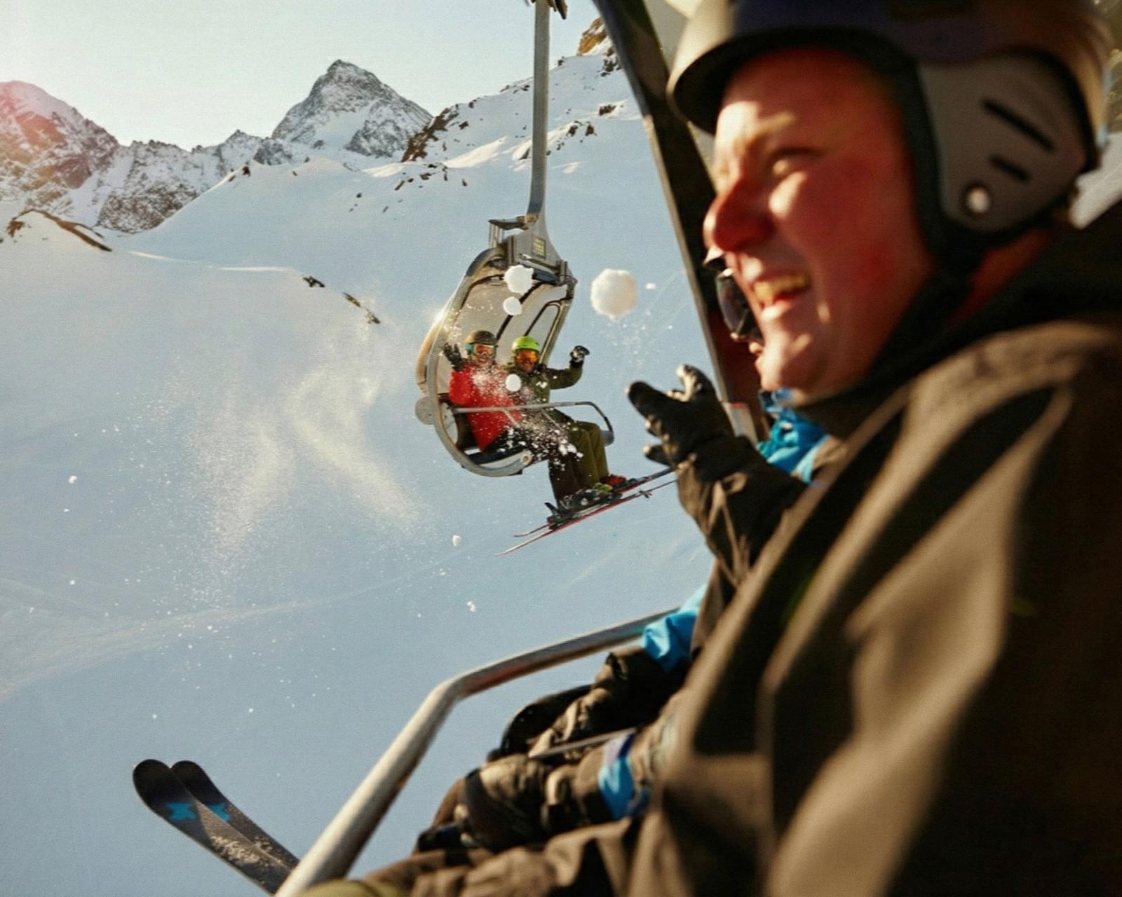A group of skiers are seen in mid-air, performing a trick on a mountain slope, with a snow-covered mountain range in the background.
