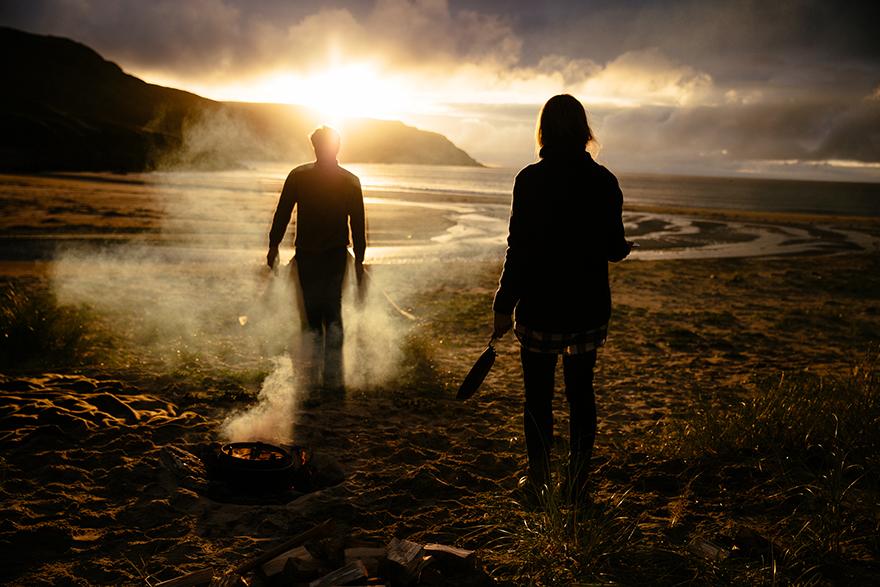 Two people walking towards a campfire on a beach at sunset, with a mountain in the background.