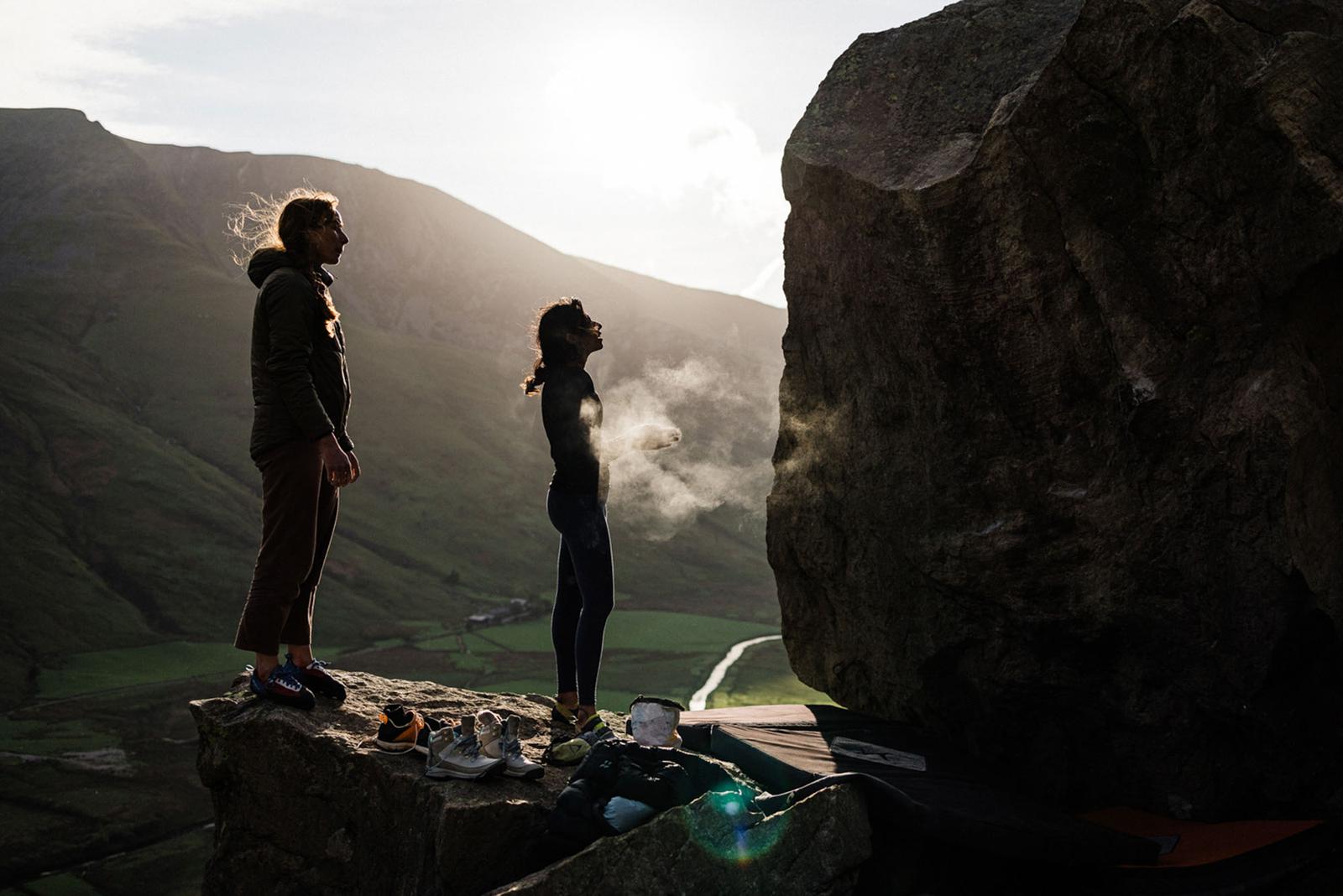 Two people standing on a rocky outcrop, looking out at a vast, mountainous landscape.
