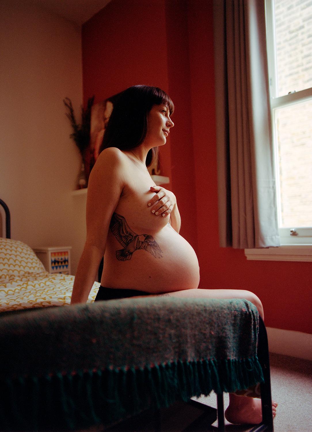 A pregnant woman with a tattoo of a bird on her abdomen, sitting on a bed, looking out the window.