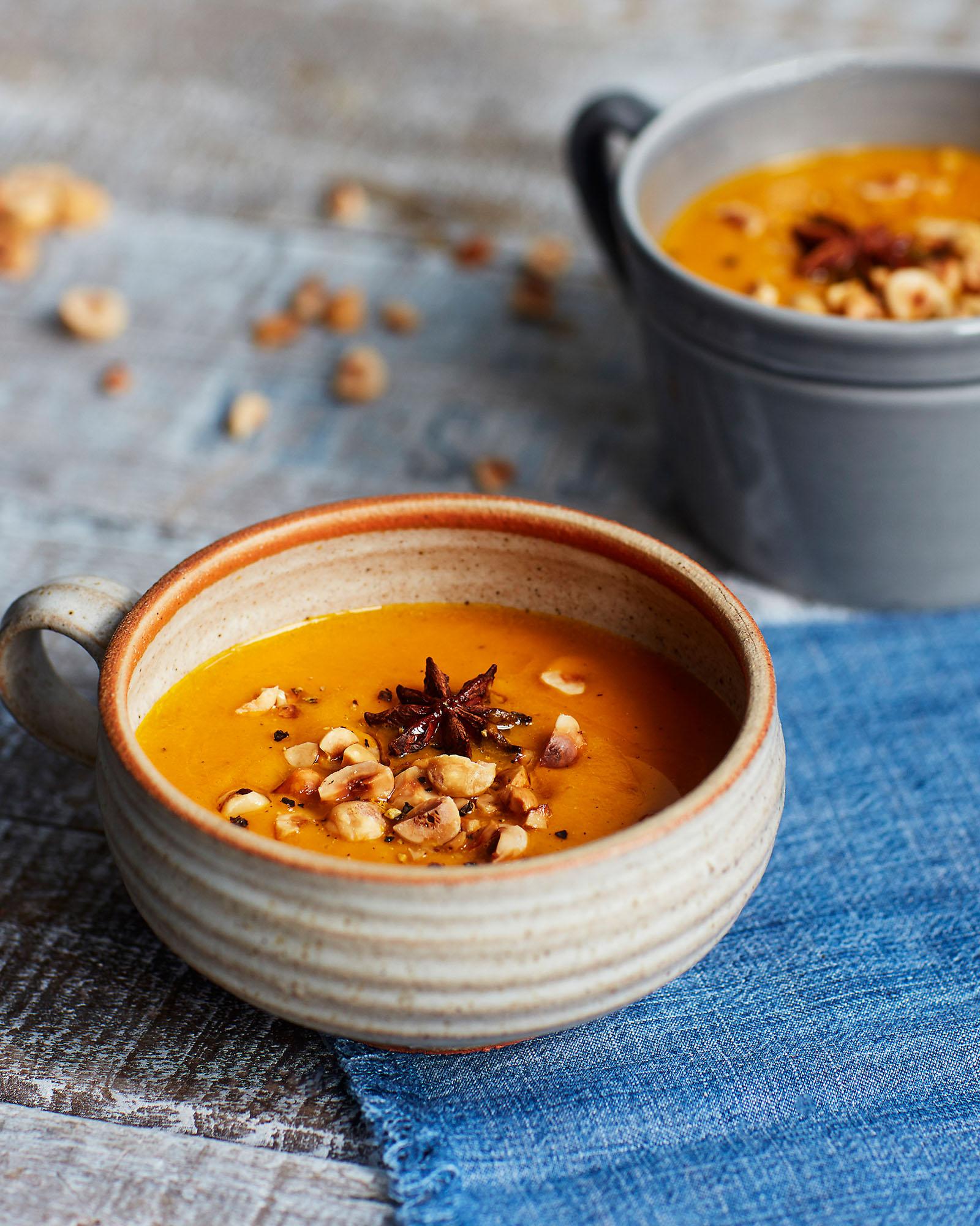 A bowl of orange soup with a star anise and hazelnuts on top, served in a white ceramic bowl with a handle, on a blue tablecloth.