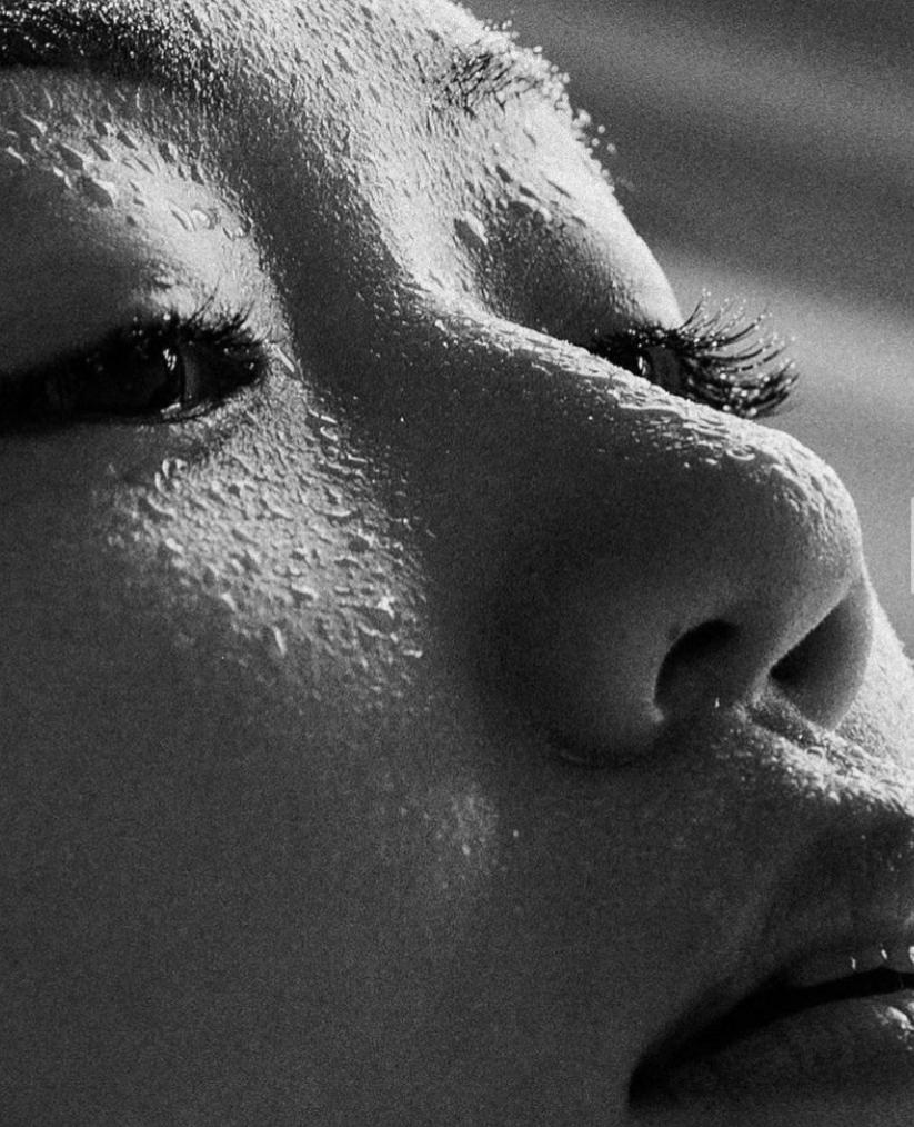A close-up of a woman's face, with water droplets on her skin and her eyes closed, as if she's just emerged from a pool or shower.