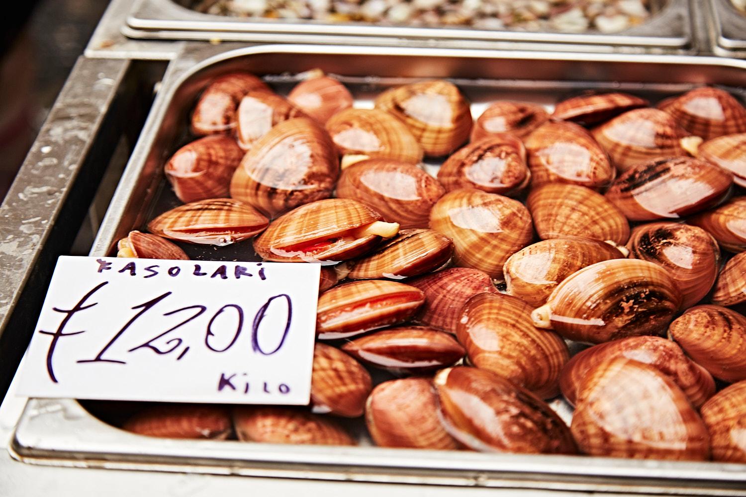A close-up of a metal tray filled with freshly cooked and steaming mussels, with a white sign in the foreground displaying the price and weight of the mussels in Italian.