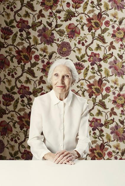 An elderly woman with white hair and a white blouse, sitting in front of a floral patterned background.