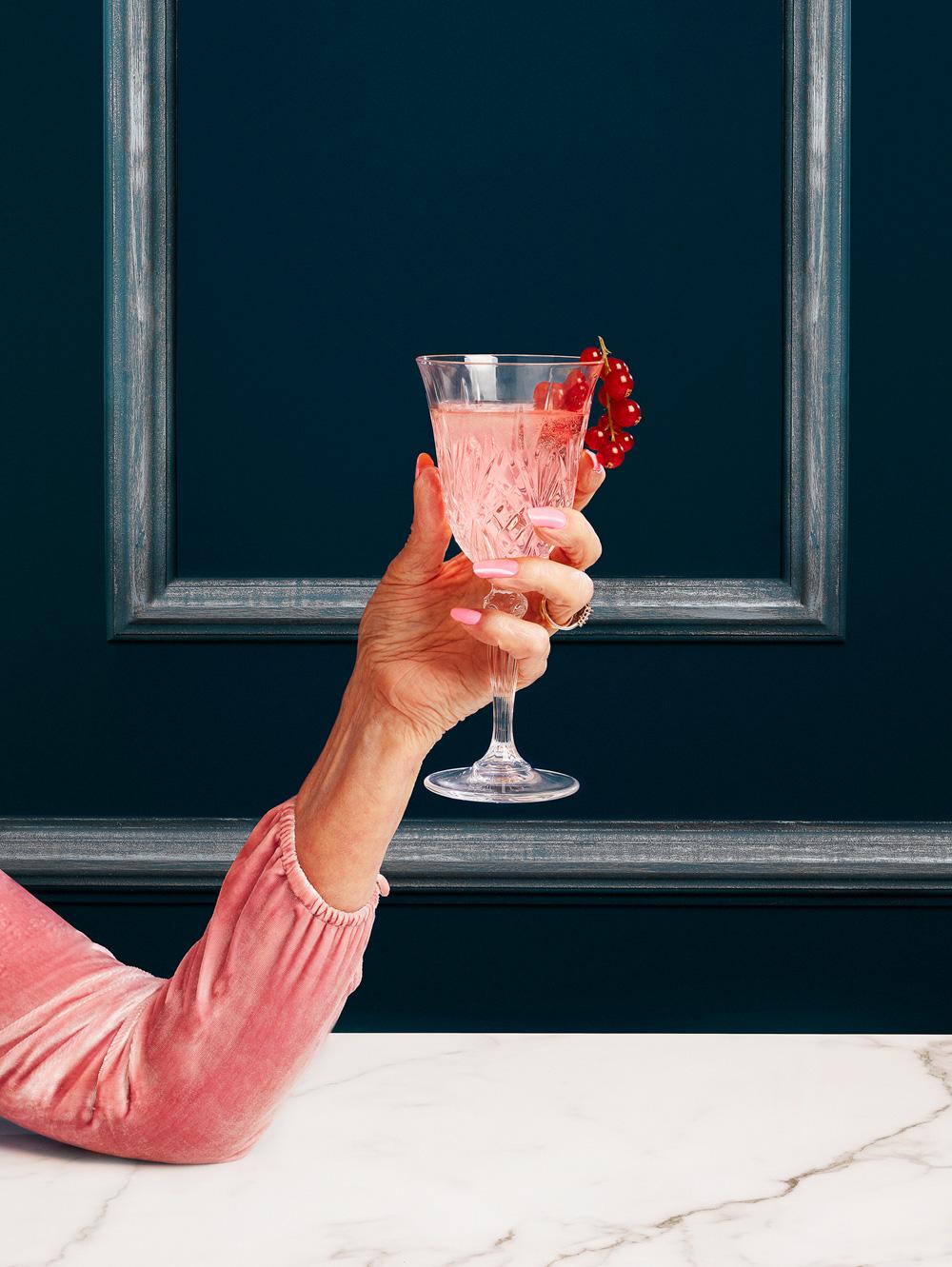 A woman's hand holding a pink cocktail in a glass, garnished with red currants, in front of a dark blue background with a large, ornate frame.