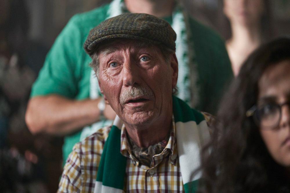 An older man with a mustache wears a flat cap, yellow and white plaid shirt, standing in front of a crowd.