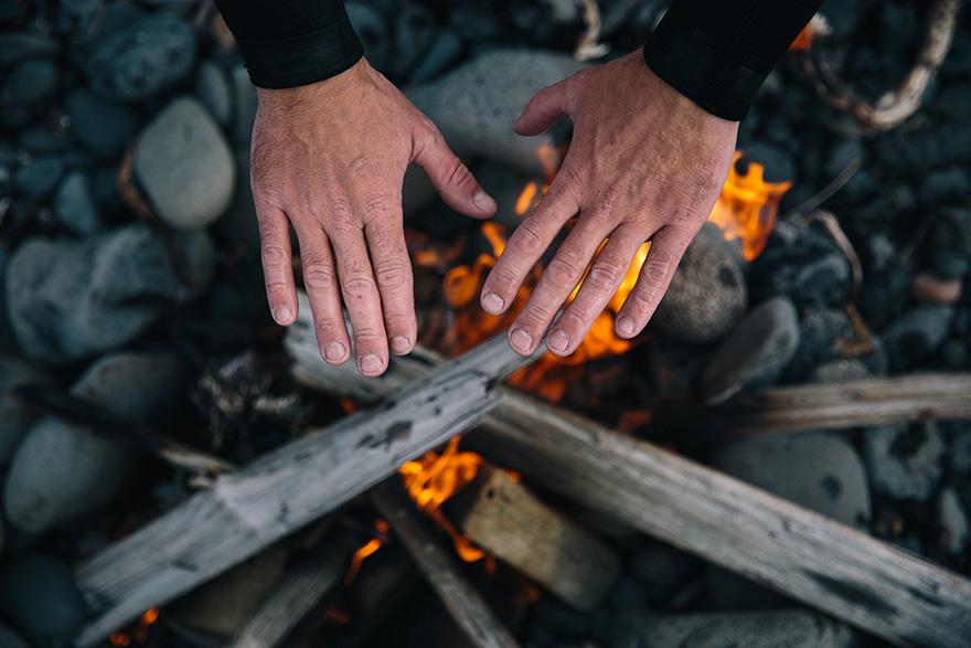 A person's hands are placed on a campfire, with flames and burning wood in the background.