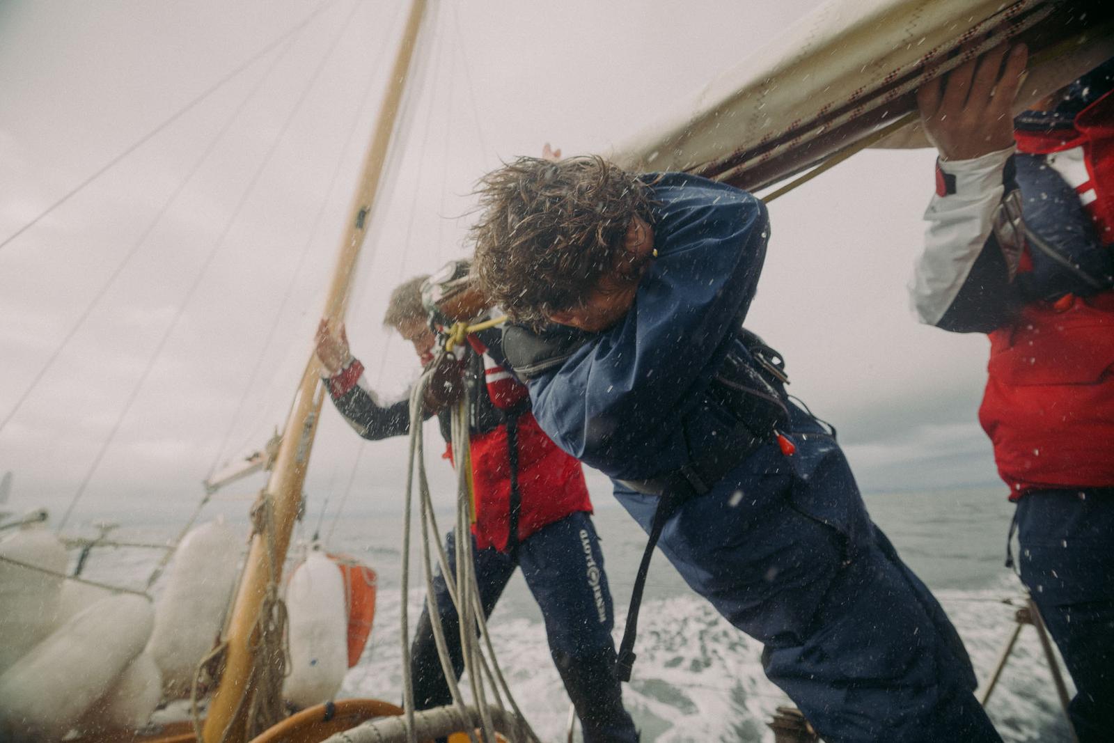 A group of people in wetsuits and life jackets are holding onto a sail on a boat in the middle of the ocean, with waves and wind creating a sense of urgency and danger.