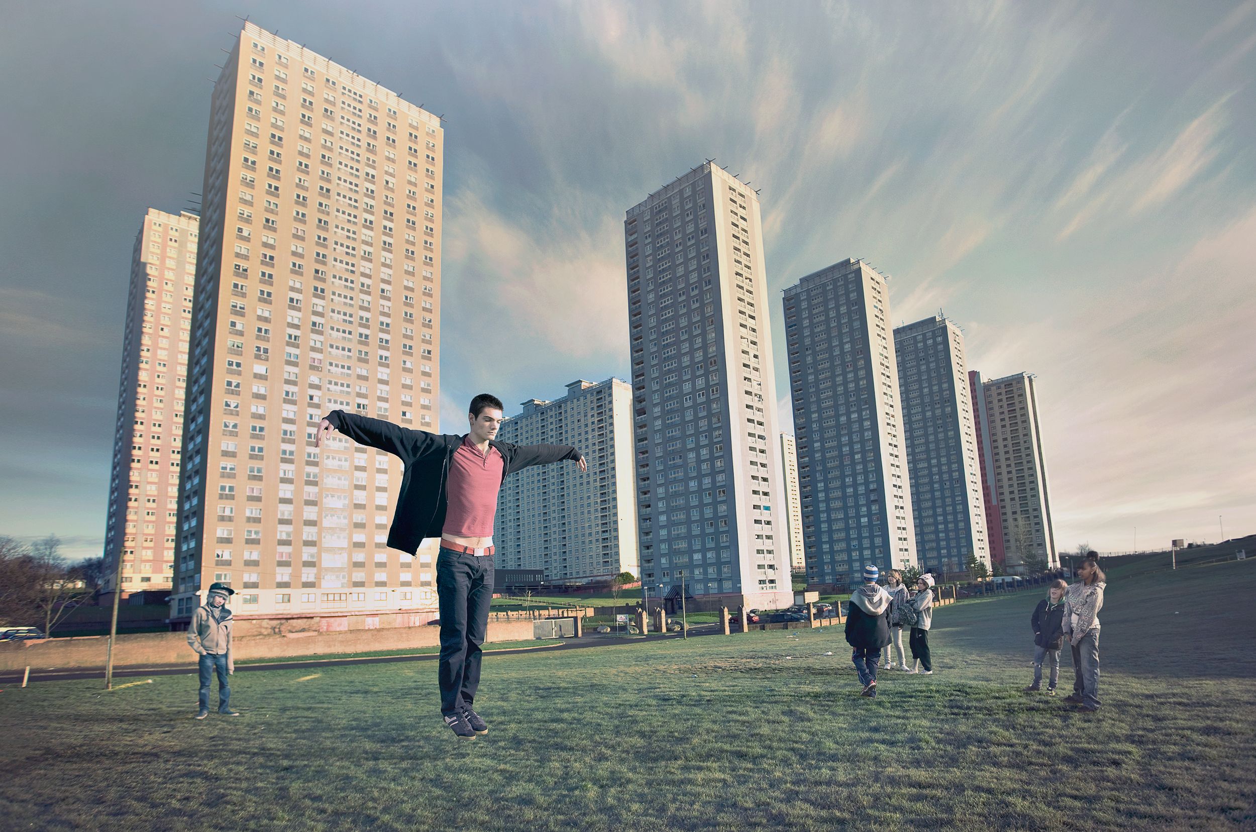 A man in a red shirt and black jacket is standing in a park with a group of children in the background, with a city skyline in the distance.