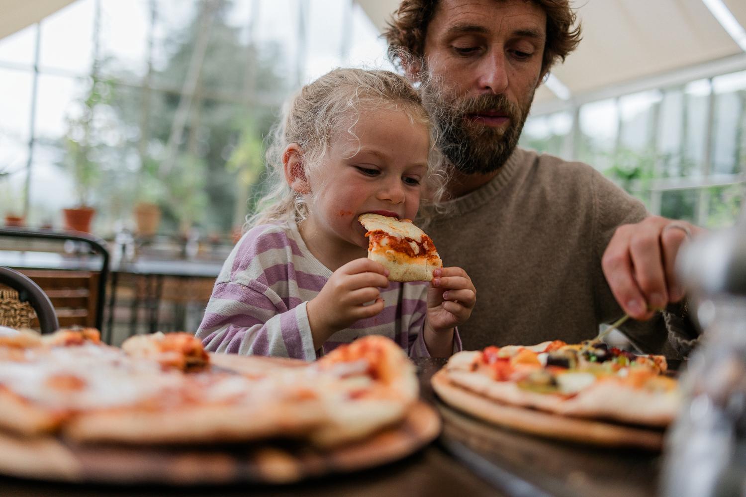 A man and a young girl eating pizza in a restaurant with a large window in the background.