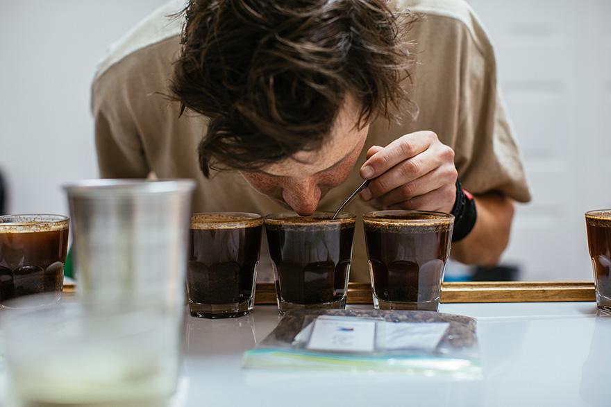 A man with a beard and a white shirt is tasting coffee from a cup.