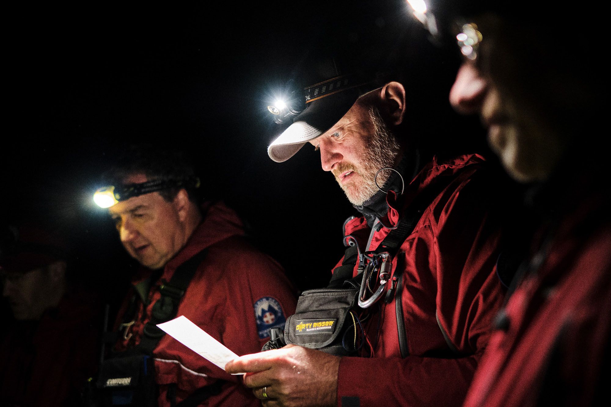 A group of people, likely a team of photographers or explorers, are huddled together in the dark, wearing red jackets and headlamps, studying a map or document.