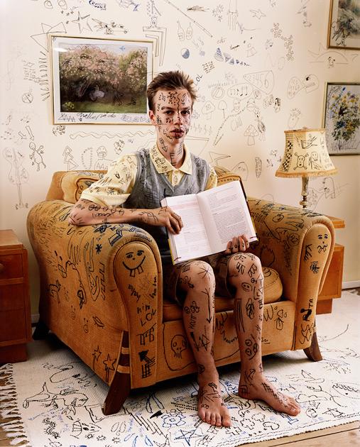 A man sits in a chair holding a book among art and art supplies.