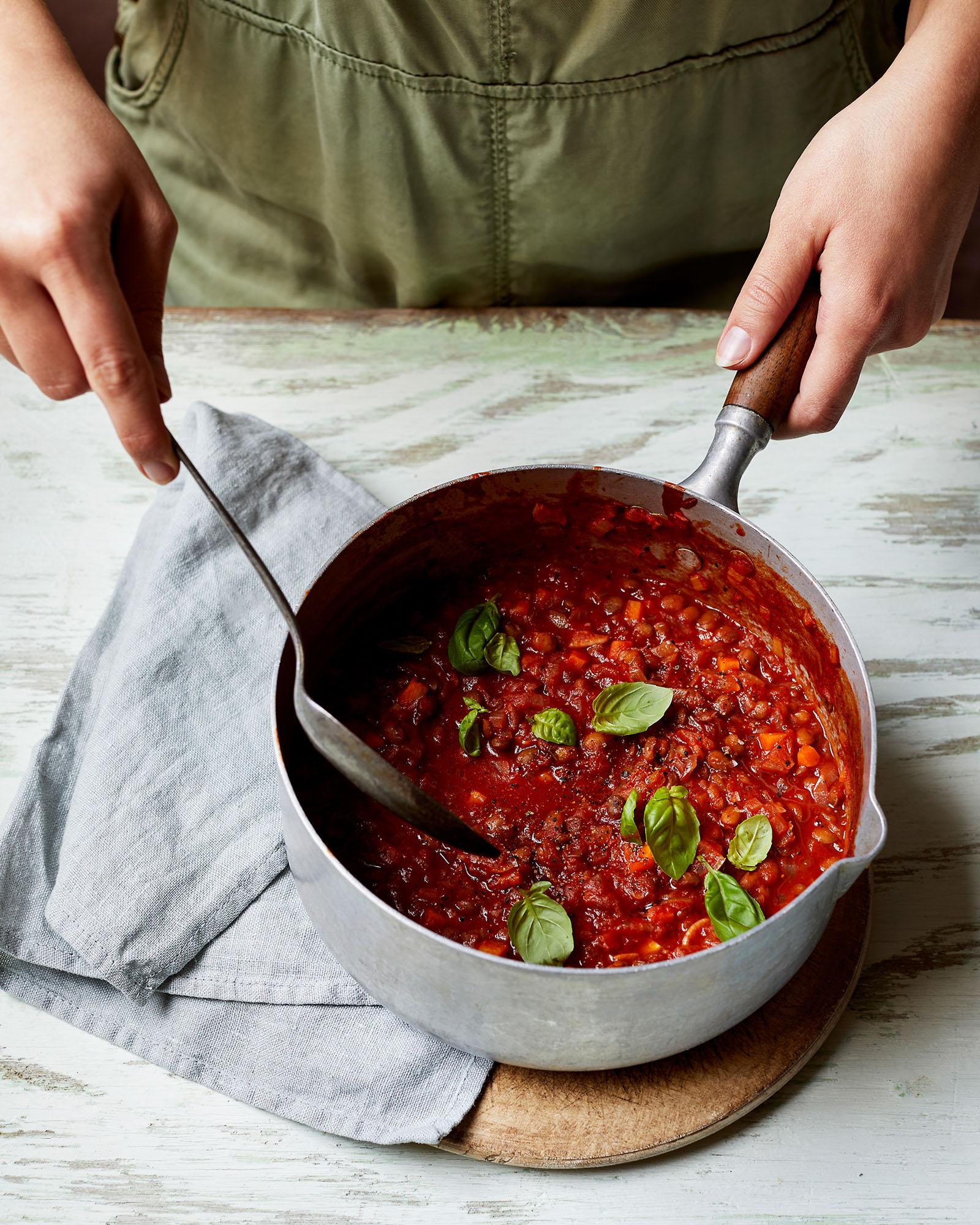 A chef's hands are stirring a large, metal saucepan filled with a rich, red tomato-based sauce, garnished with fresh basil leaves.