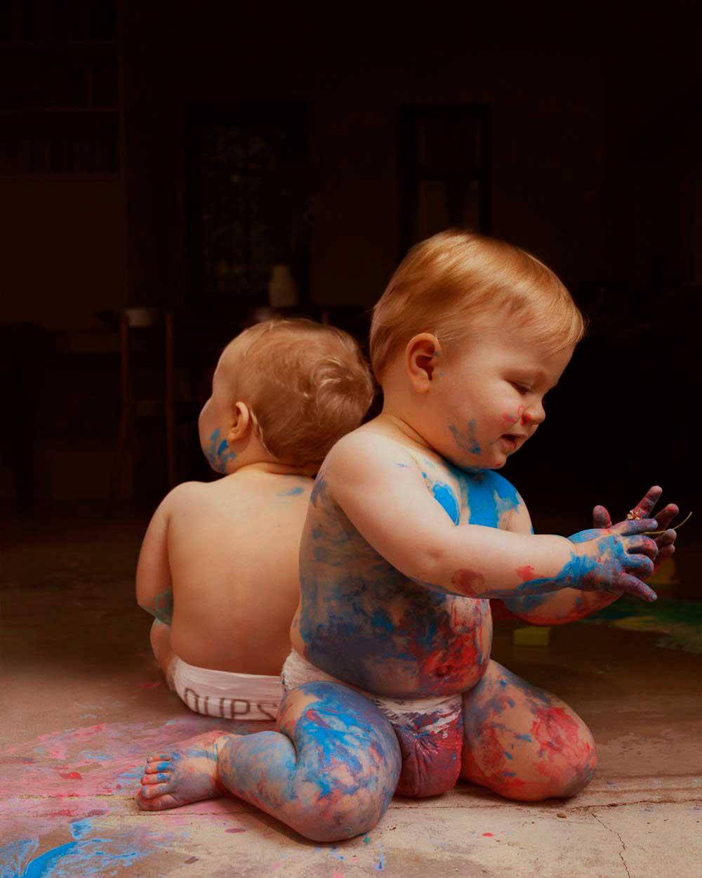 Two toddlers, both male, covered in paint, sitting on the floor, wearing only diapers.