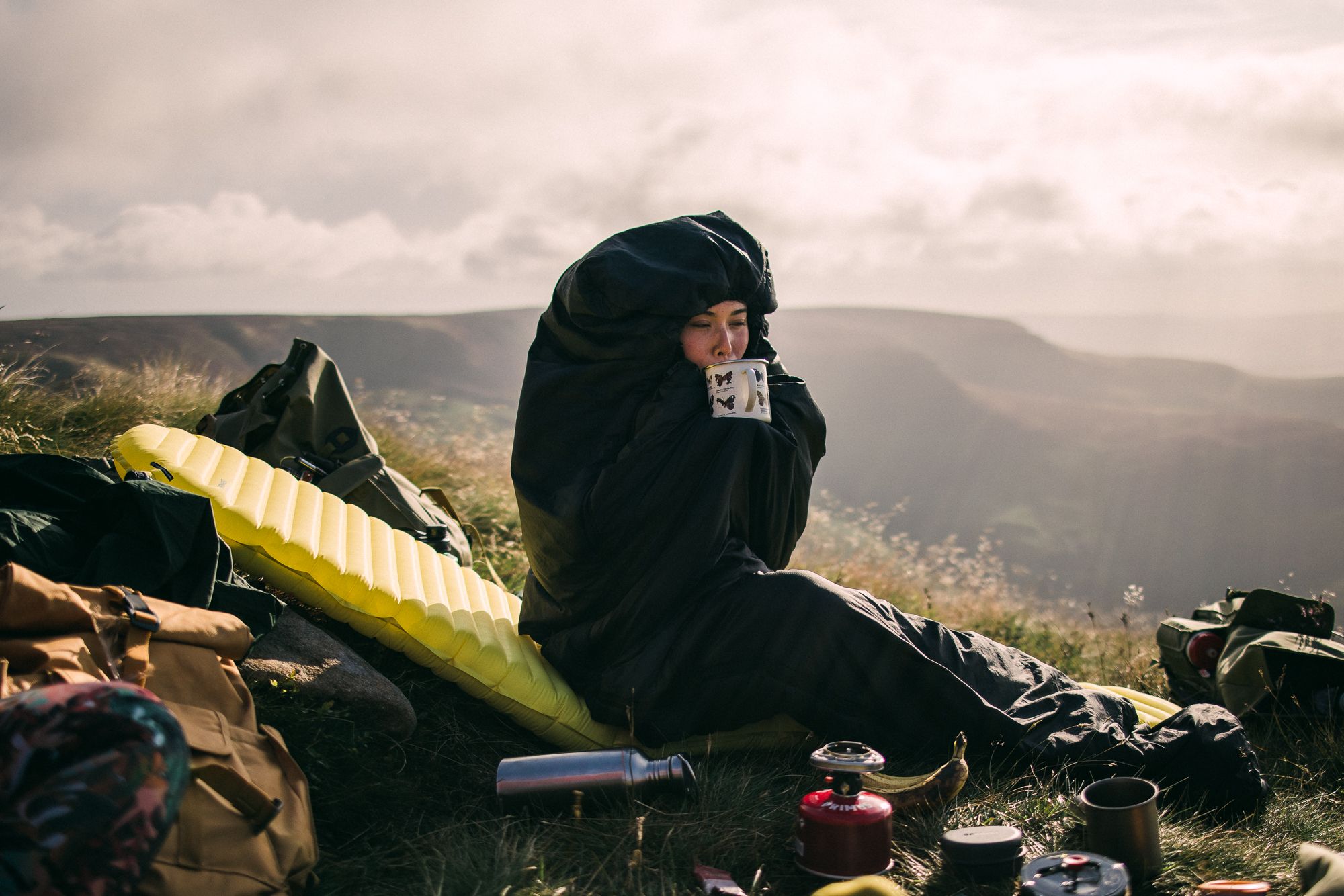 A woman in a black sleeping bag and a black and yellow sleeping mat sits on a rock, holding a cup of coffee in a scenic, mountainous landscape.