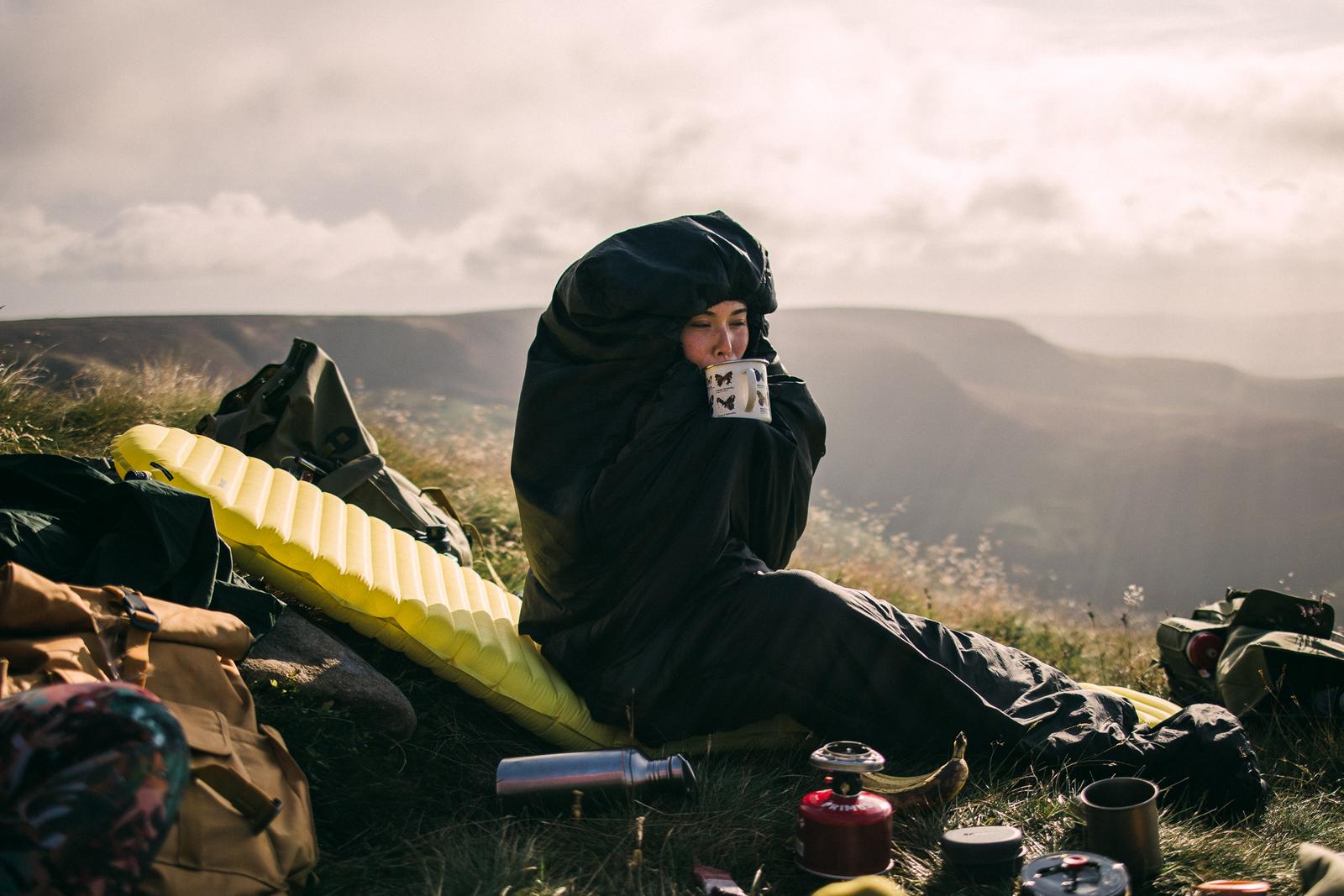 A woman in a black sleeping bag and a black and yellow sleeping mat sits on a rock, holding a cup of coffee in a scenic, mountainous landscape.