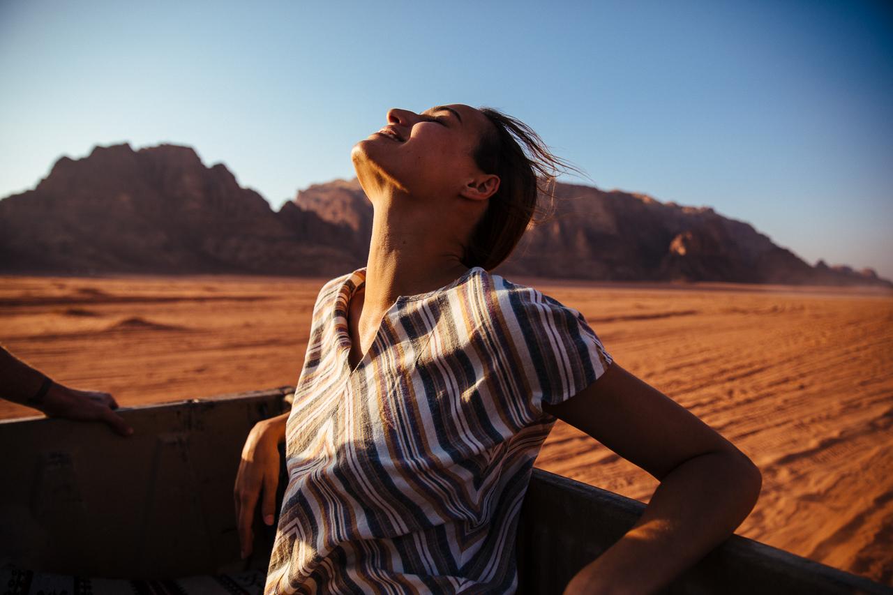 A woman in a striped shirt is sitting in the back of a truck in the desert, with her head thrown back and her eyes closed, enjoying the sun on her face.
