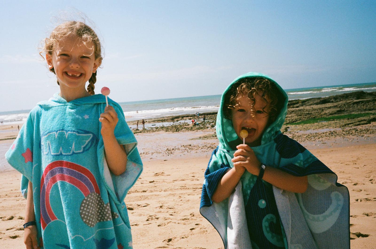Two children, one with a lollipop, stand on a beach, smiling at the camera. The child on the left has a blue towel with a rainbow design.