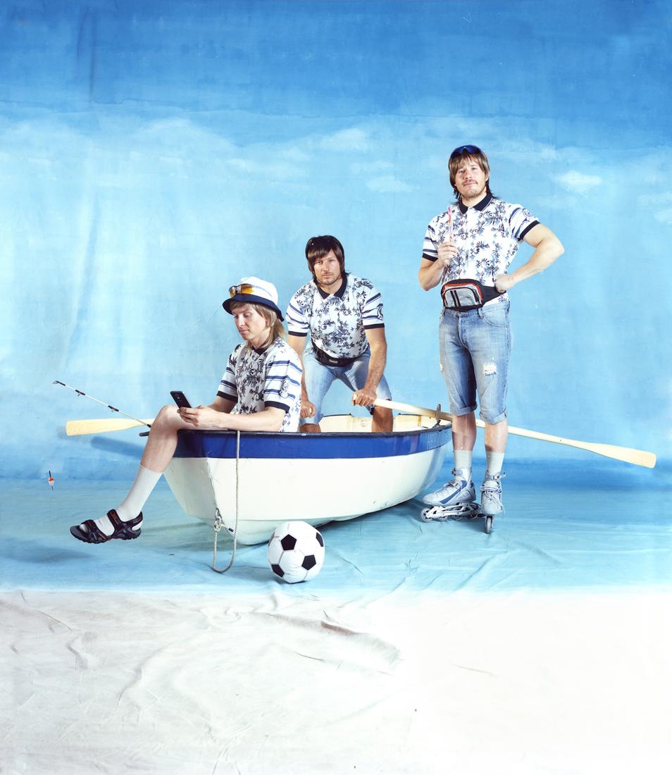 Three men in a small, white and blue boat, one of them holding a smartphone, with a soccer ball and a fishing rod in the background.