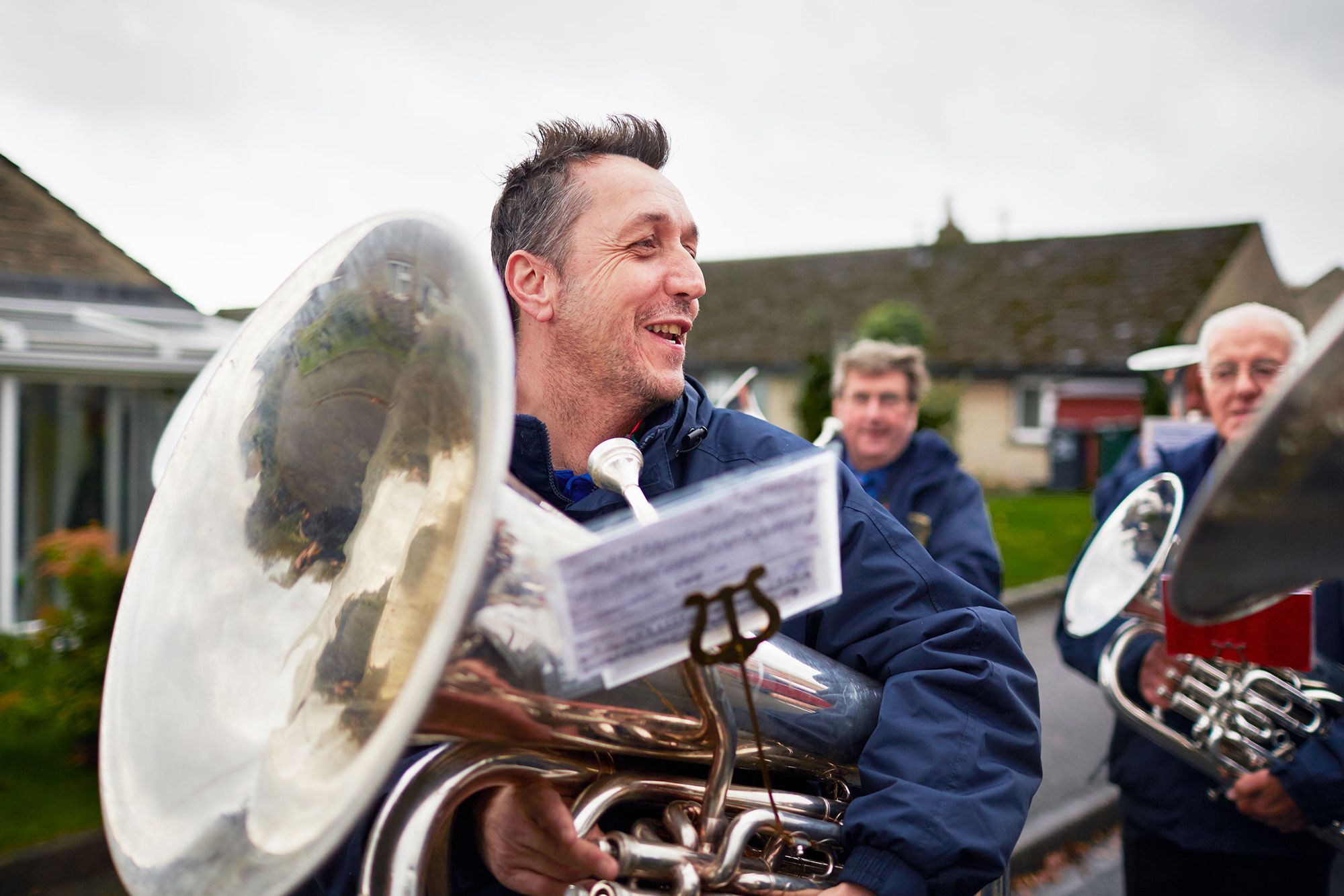 A group of men, likely a brass band, marching in a parade or street performance, wearing blue jackets and holding brass instruments, including a large tuba.
