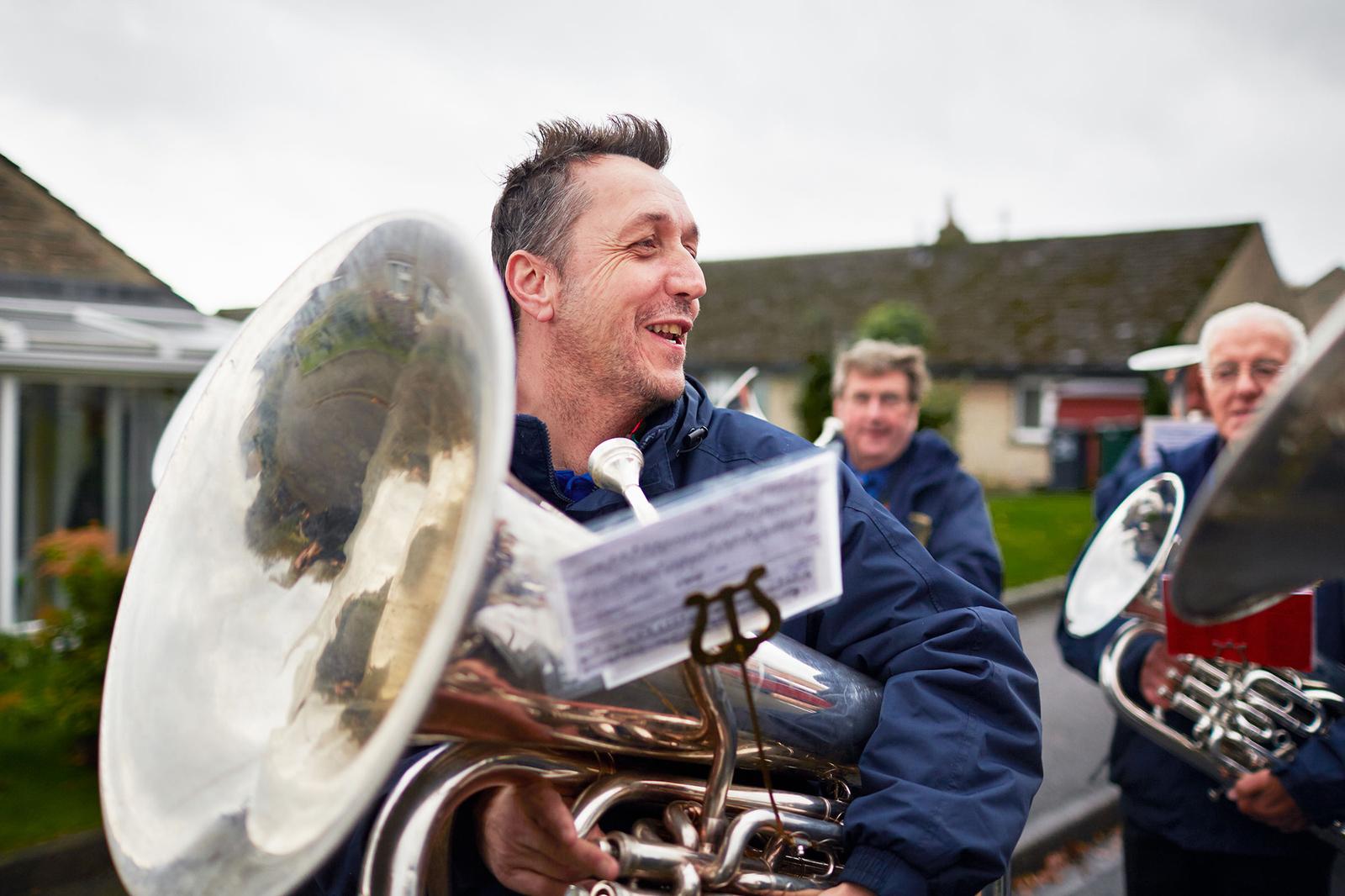 A group of men, likely a brass band, marching in a parade or street performance, wearing blue jackets and holding brass instruments, including a large tuba.