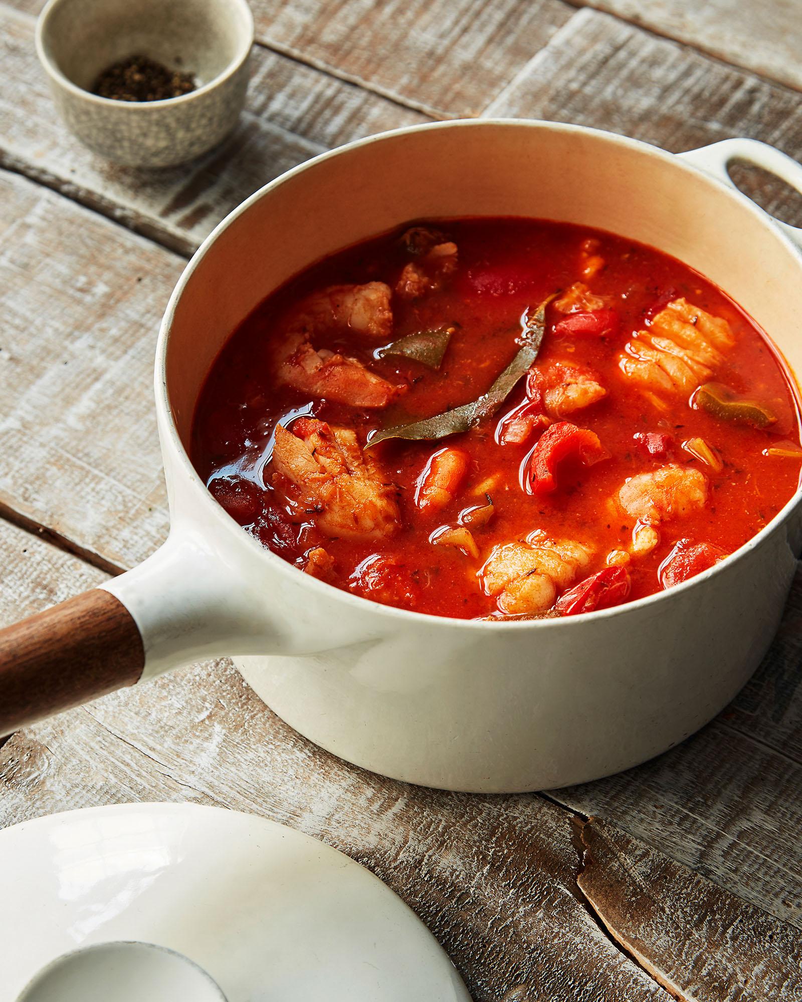 A large, white, ceramic pot filled with a rich, red, stew-like soup, with chunks of meat and vegetables visible.