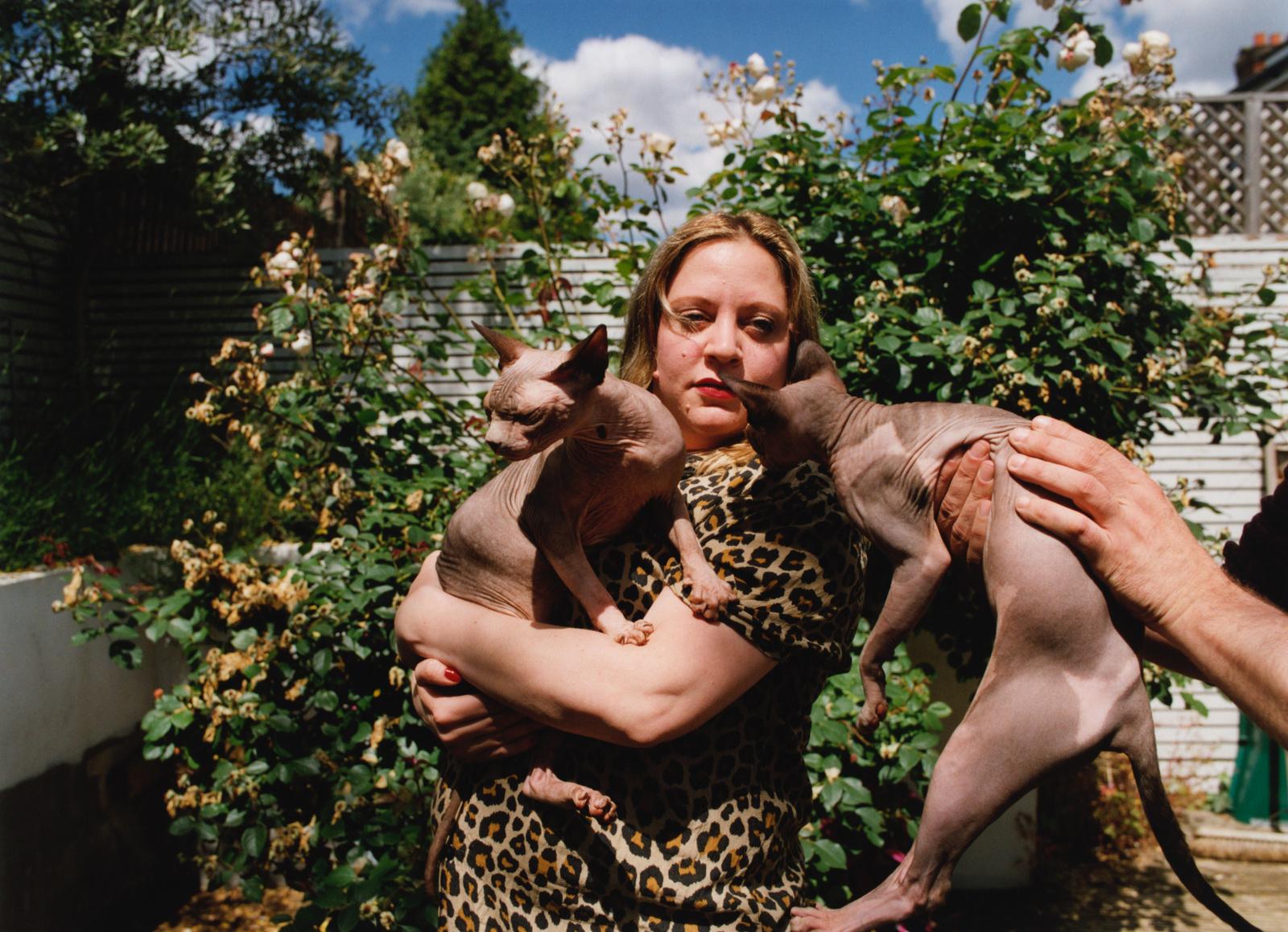 A woman in a leopard-print dress holding two hairless cats in her arms, in a garden or outdoor setting.