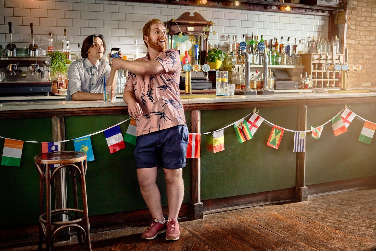 A man in a bar, holding a beer and taking a selfie with a bartender in the background.