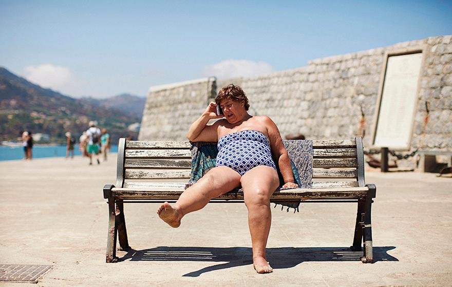 A woman in a blue and white swimsuit is sitting on a bench, talking on her phone.