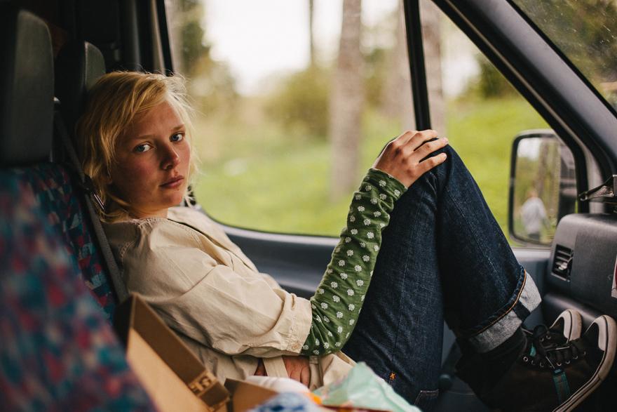 A young woman with blonde hair and a white shirt is sitting in the driver's seat of a car, looking directly at the camera with a neutral expression.