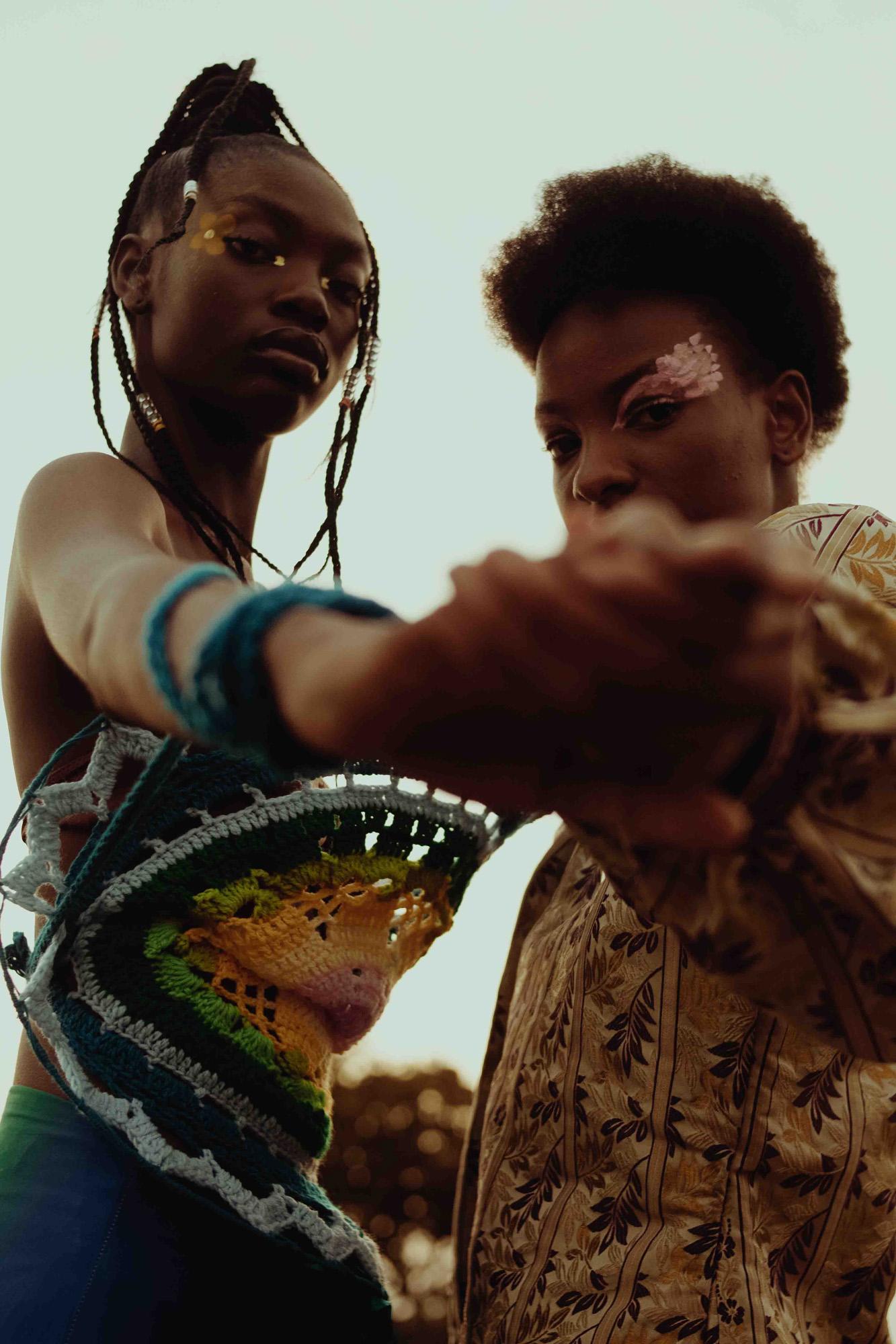 A woman and a man in traditional African clothing stand together, both with Afro hairstyles, holding their respective garments, against a blurred outdoor backdrop.
