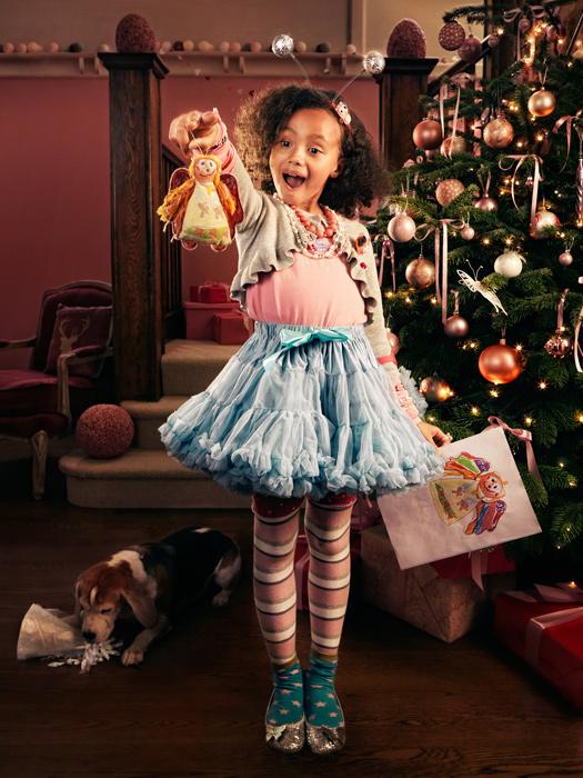 A young girl holds a doll in front of a Christmas tree with gifts and a dog.
