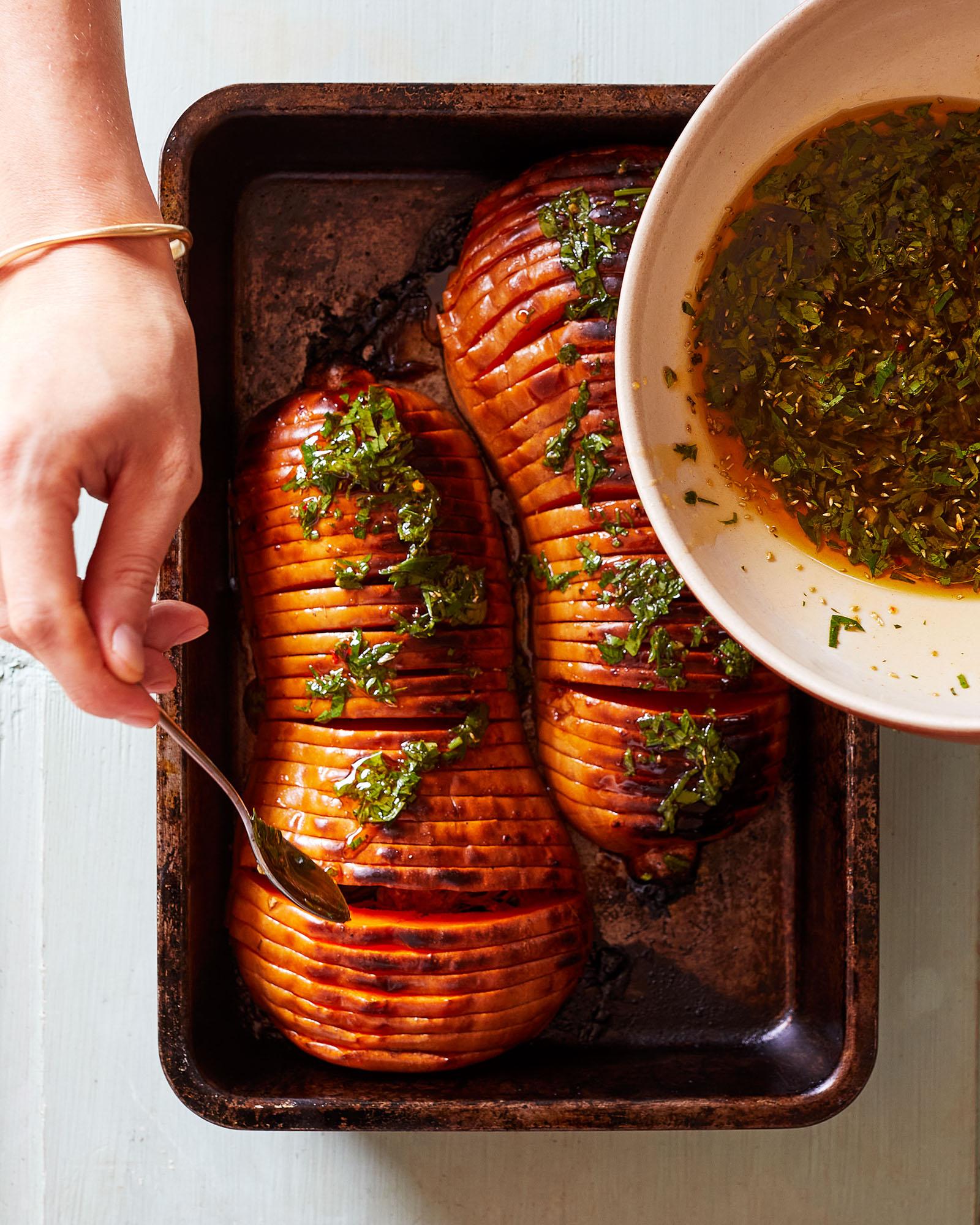A person's hand is holding a metal spoon over a baking dish containing two large, roasted, spiral-cut sweet potatoes, drizzled with a brown sauce and garnished with chopped herbs.