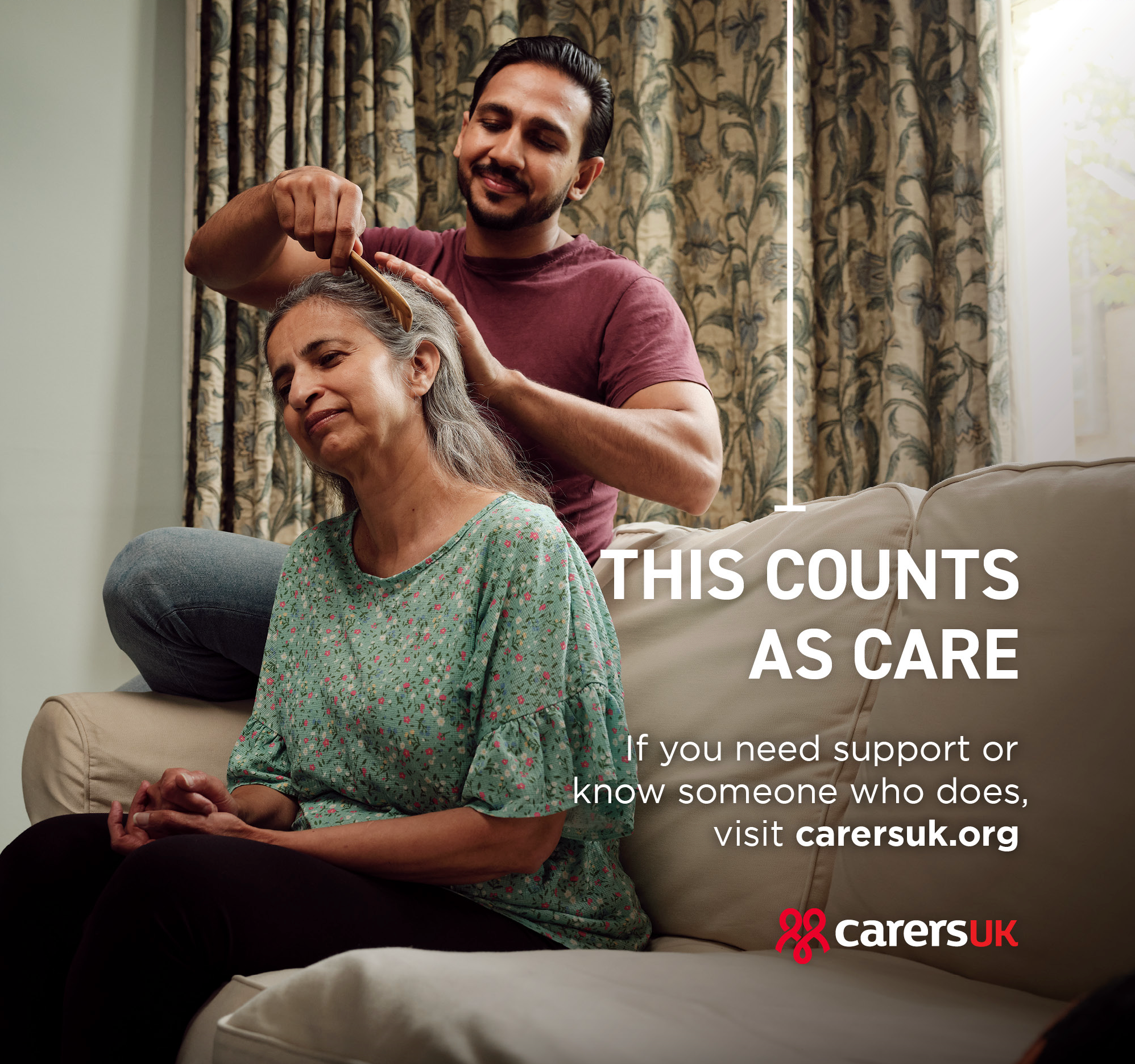 A man is sitting on a couch, brushing the hair of an older woman who is sitting on the couch in front of him.
