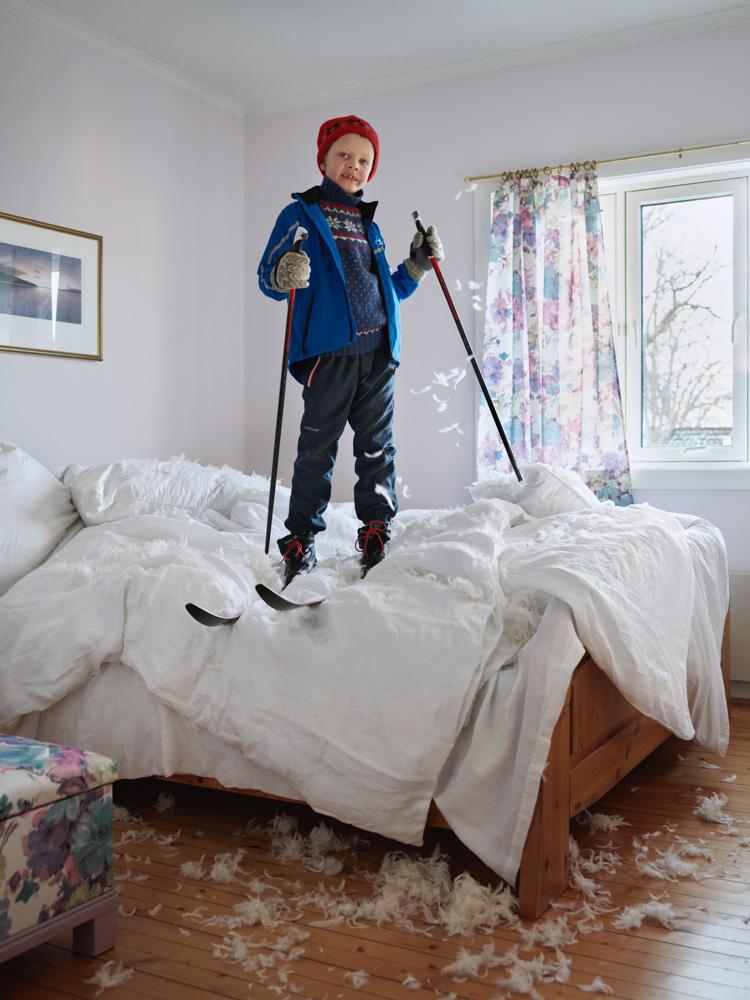 A young boy in a blue jacket, red hat, and snow gear is standing on a bed, surrounded by feathers and snow gear, in a bedroom with a window and a tree outside.