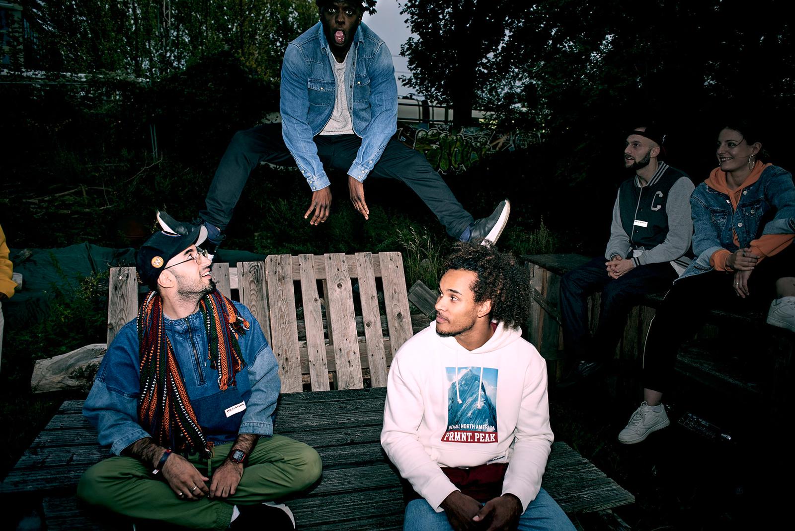 A group of people sitting and standing on a wooden bench in a park with a relaxed atmosphere.