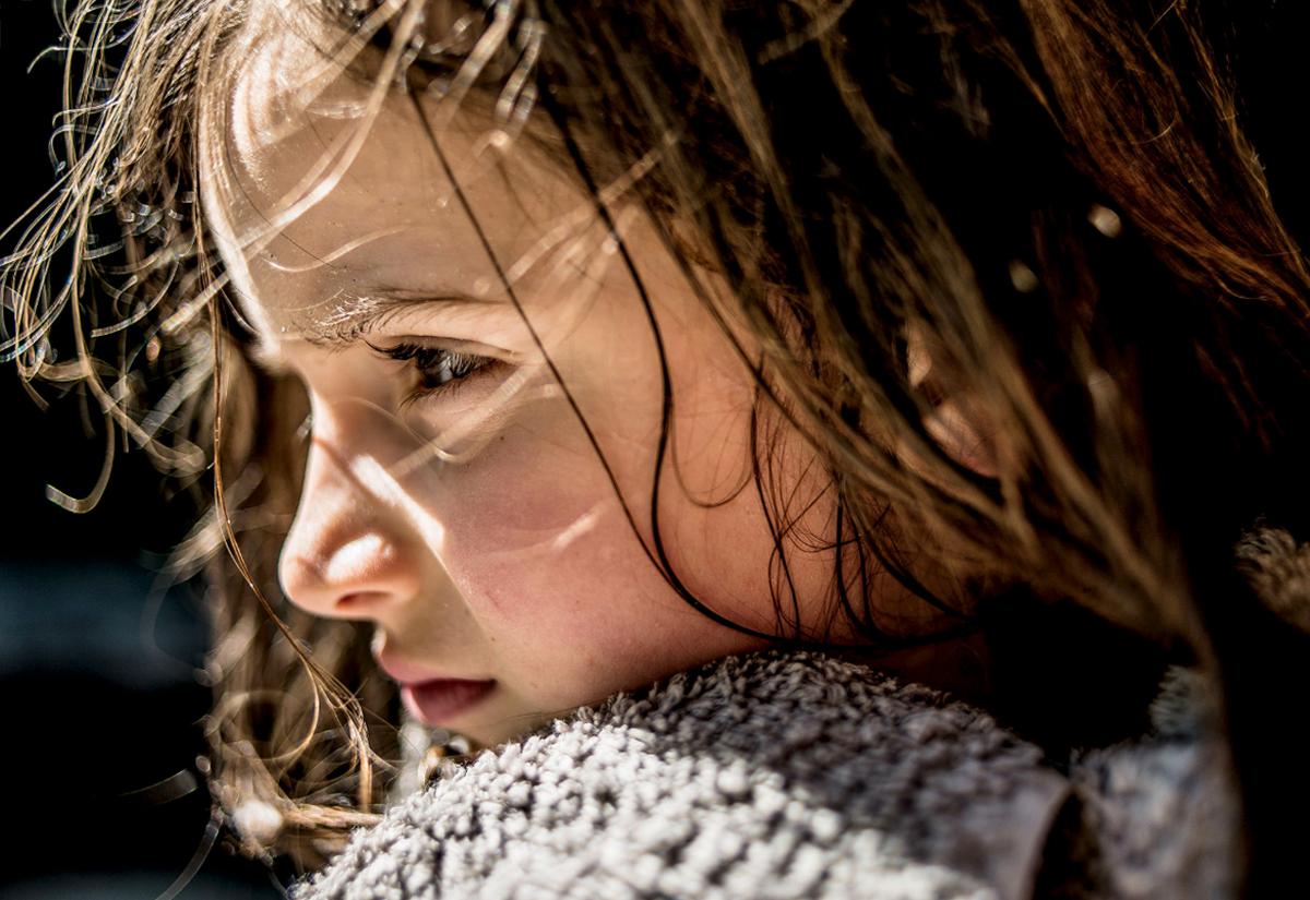 A young girl with wet hair and a towel around her neck, looking out of a window with a mix of emotions on her face.