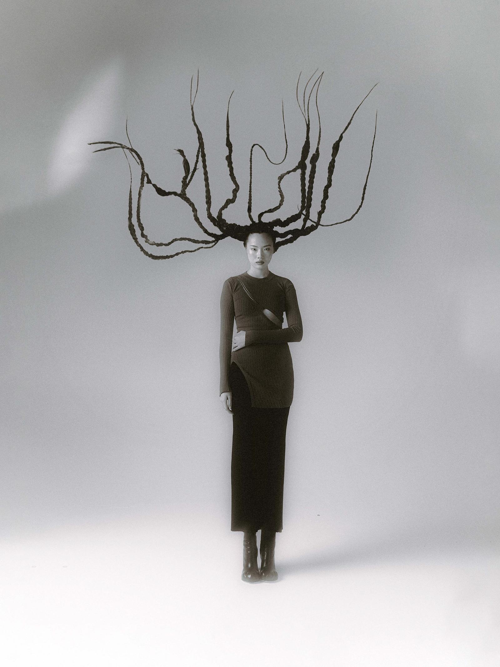 A woman with long, thin, black hair styled in multiple braids, wearing a black dress and standing in a minimalist, white background.