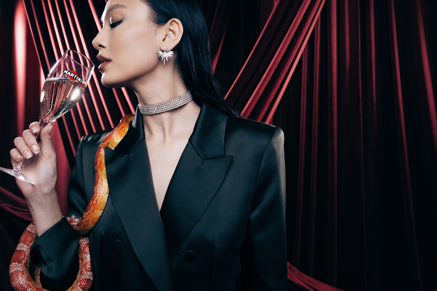 A woman holds a glass of champagne in a luxurious setting with a snake wrapped around her.