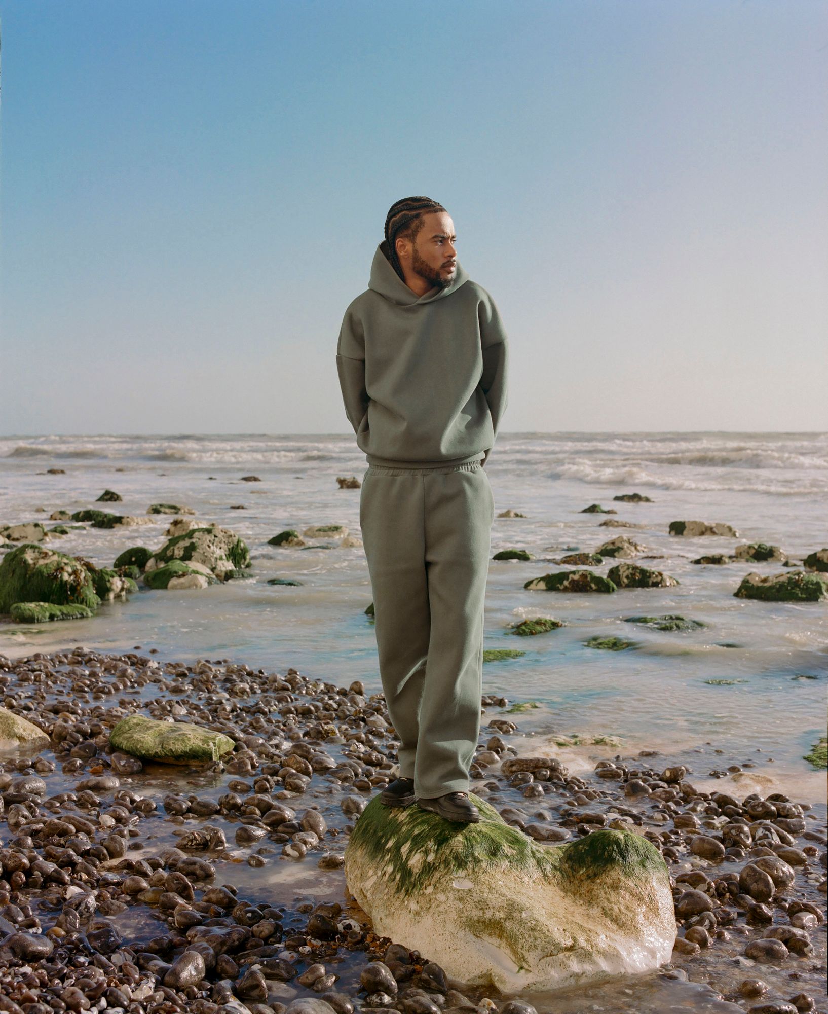 A man standing on a rock at the beach, wearing a green hoodie and sweatpants.