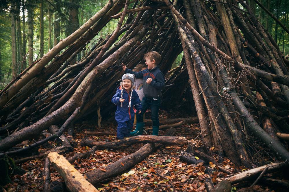 Two children in blue rain gear stand in front of a large, rustic structure made of logs and branches in a forest. They appear to be enjoying themselves, with one child looking.